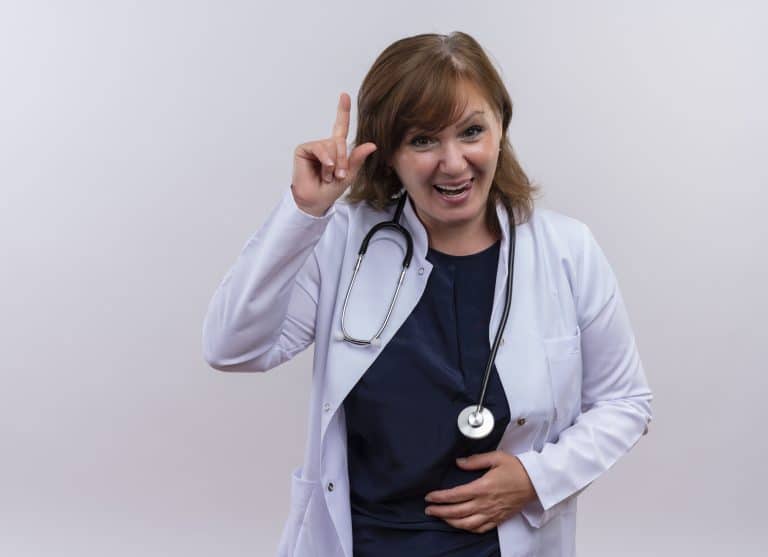 joyful middle-aged woman doctor wearing medical robe and stethoscope pointing with finger up and putting hand on belly on isolated white background with copy space